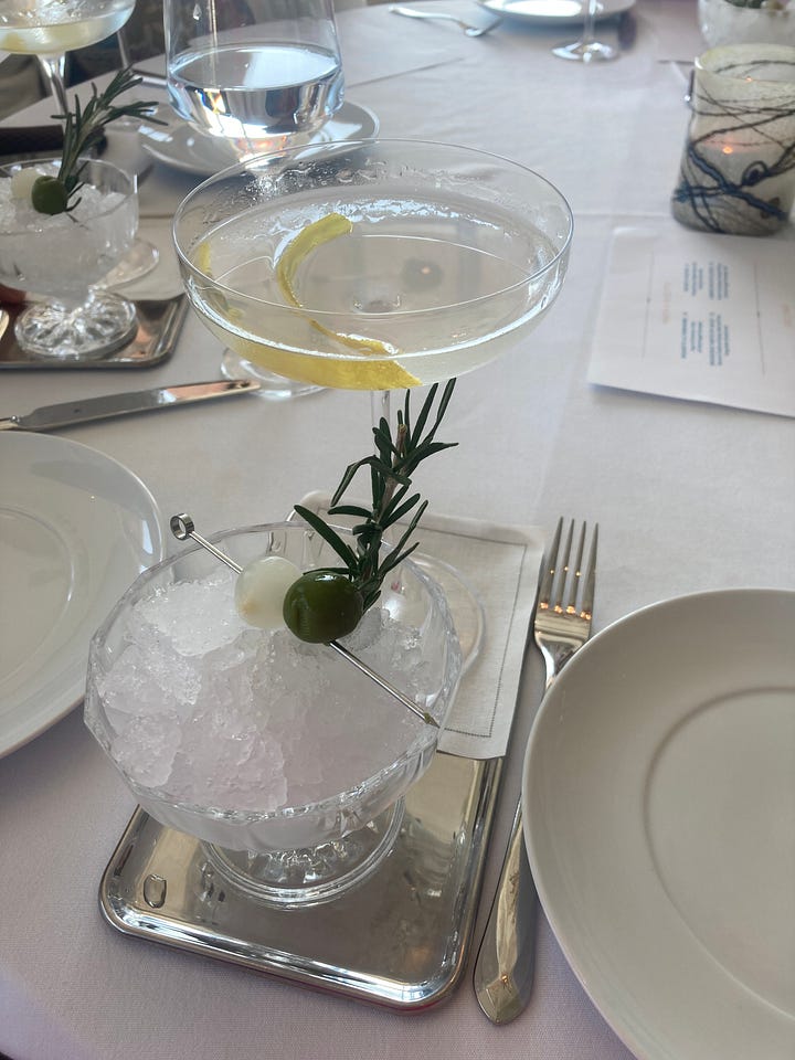 Photo 1: Vicky and Charlie standing on a balcony at sunset. Charlie wears a blue suit and pink tie and holds a champagne flute. Vicky wears a hot pink dress. Photo 2: The Capolinea martini, a gin martini with lemon with optional olive and rosemary over ice on the side. Photo 3: The old-fashioned bar in the entryway of Capolinea with fully stocked brass shelves in back. Photo 4: The layout of Capolinea. The rollable martini cart is in the foreground with a pot of pink flowers next to it. Round tables and chairs stretch all the way to the back of the restaurant through a series of light green arches.