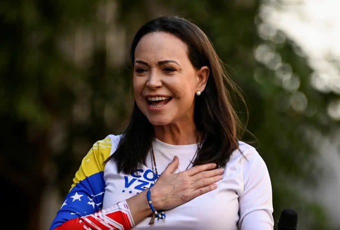 Woman with shoulder-length dark hair smiles broadly outdoors in natural light, wearing white long-sleeve shirt with blue-yellow-red Venezuelan flag stripes on left sleeve, hand over heart, bracelet on wrist, earrings visible, green foliage background.