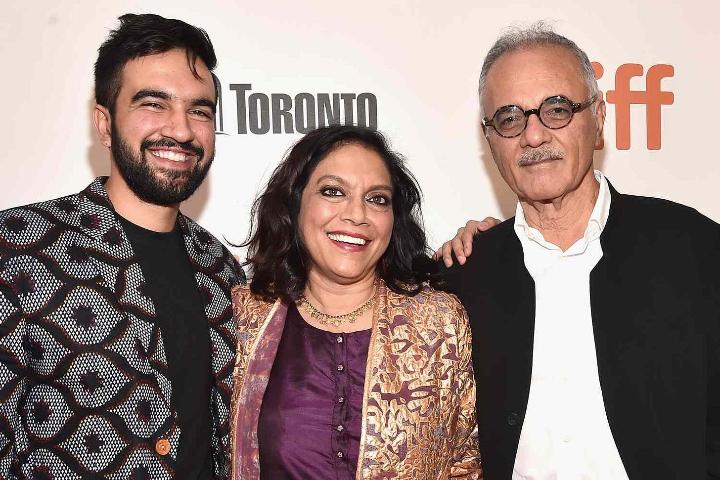 A photo of Zohran Mamdani, with a full beard, black hair and a big smile, alongside his parents. His mom has a fancy purple shirt with gold jacket on and has brown skin and black shoulder length hair. His dad has short grey hair, round black rimmed glasses on and a black jacket.