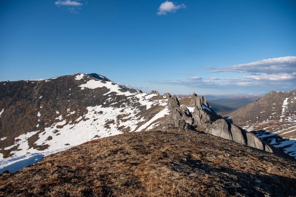 A row of jagged granite spires runs along a snow-streaked ridge, with a tundra foreground and a long valley opening to faint distant ranges under a deep blue sky.