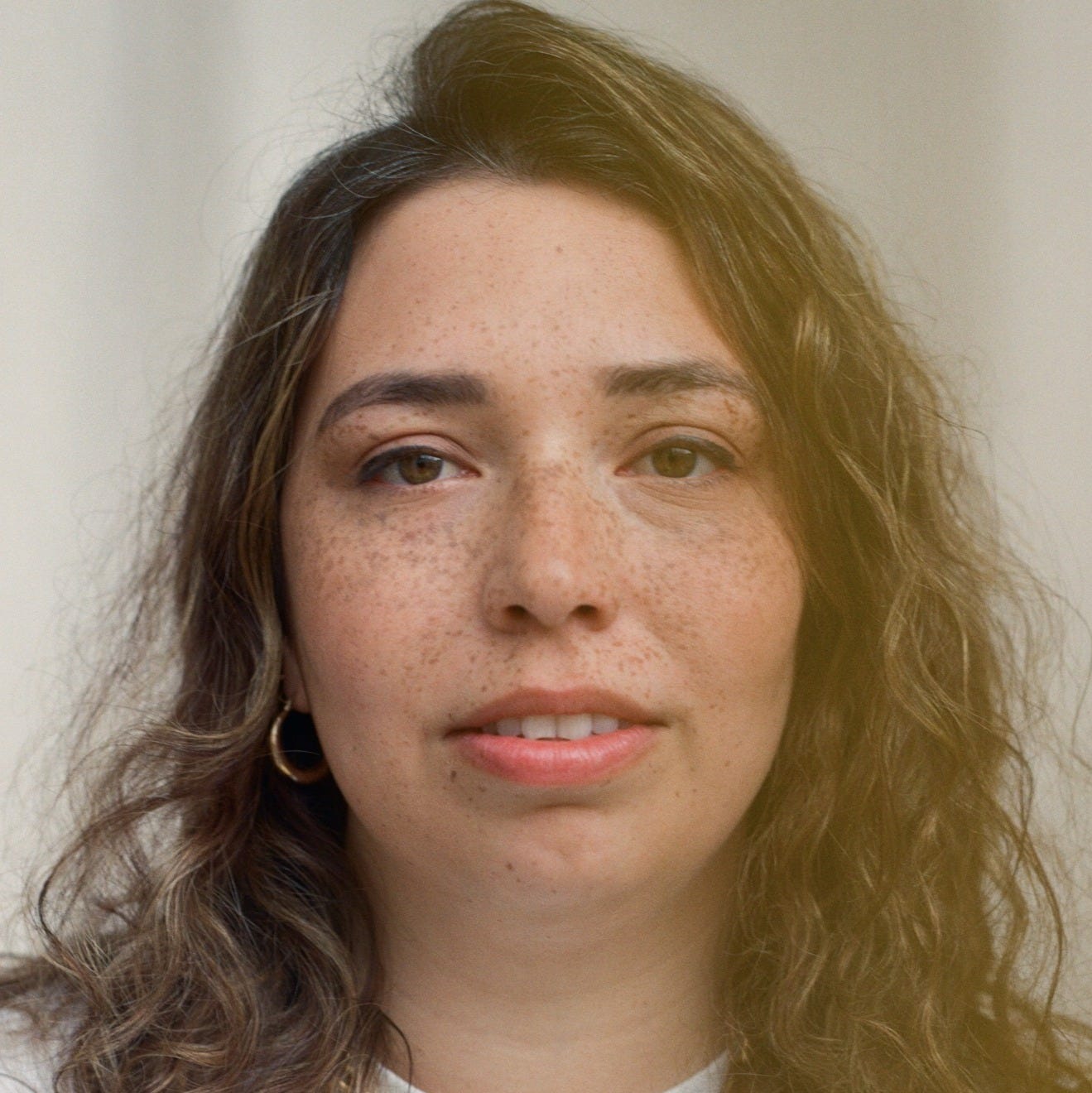 Portrait of writer Emma van Straaten, a British-Mauritian novelist, photographed by Robin Christian, showing her with wavy brown hair, light freckles, gold hoop earrings, and a gold chain necklace, wearing a white top against a soft neutral background, used for her interview with Finbarre Snarey from Tarot DMs. Portrait of writer Emma van Straaten, a British-Mauritian novelist, photographed by Robin Christian, showing her with wavy brown hair, light freckles, gold hoop earrings, and a gold chain necklace, wearing a white top against a soft neutral background, used for her interview with Finbarre Snarey from Tarot DMs.