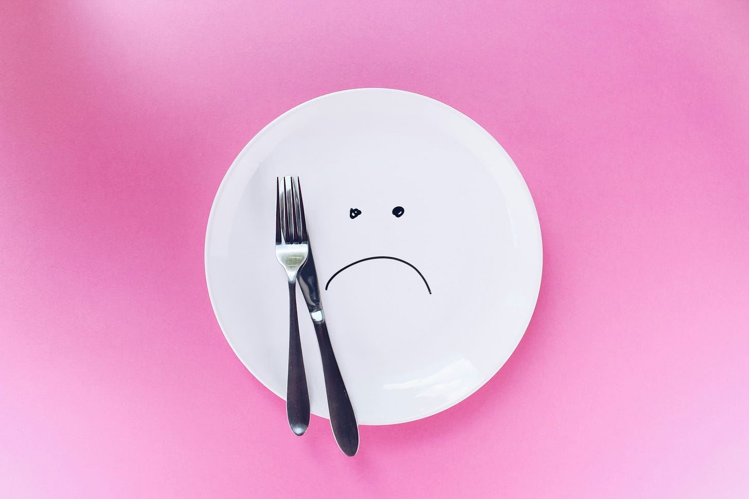 a white plate with a sad face is on a pink background. An unused fork and knife rest on the plate