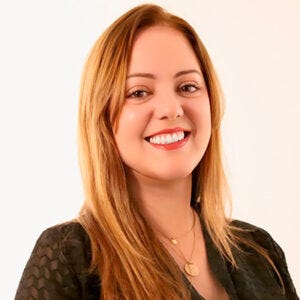 A imagem mostra uma pessoa de frente, enquadrada do peito para cima, com fundo claro e neutro. Ela tem pele clara e cabelos longos, lisos, de tom loiro escuro ou castanho claro, repartidos de lado e caindo sobre os ombros. O rosto é oval, com sobrancelhas bem definidas e olhos claros.Ela sorri amplamente, mostrando os dentes, transmitindo uma expressão acolhedora e confiante. Usa maquiagem leve, com destaque para o batom em tom rosado ou avermelhado. Veste uma blusa ou vestido escuro, possivelmente preto, com textura discreta no tecido. No pescoço, usa um colar delicado com pingentes pequenos e circulares. A postura é ereta e relaxada, e a iluminação é suave, deixando a imagem clara e profissional.