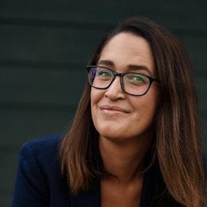 The cropped portrait shows the woman from the shoulders up, angled slightly toward the camera. Her glasses, face, and dark blazer are in focus, while the muted dark green background remains plain and unobtrusive.