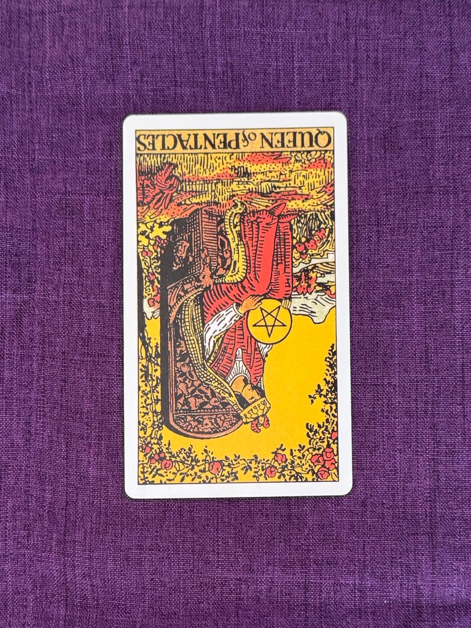 Tarot card upside down shows: There’s a woman sitting on a stone throne decorated with carvings of fruit trees, goats, angels among other symbols. ,She has a crown and holds a coin with a star in the middle on her lap. Plants and flowers surround her, a rabbit jumps near her.