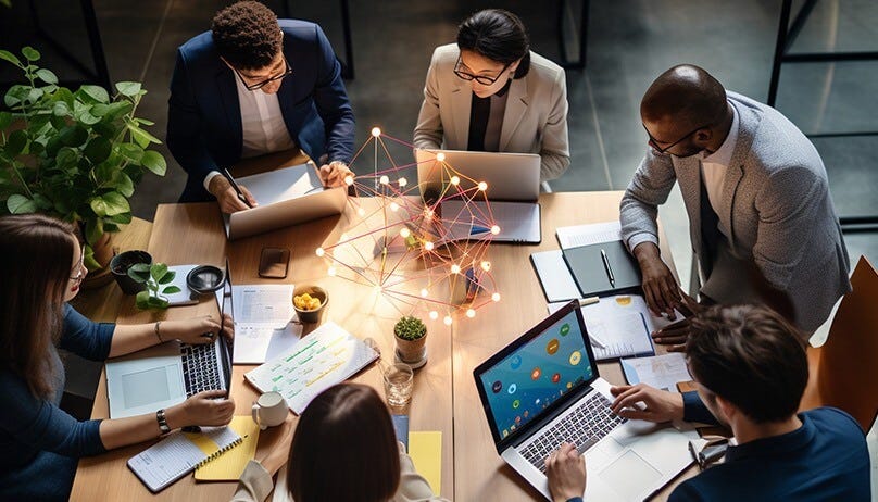 A group of six diverse professionals sits around a table working collaboratively on laptops with papers and office supplies, with a glowing geometric structure in the center. A group of six diverse professionals sits around a table working collaboratively on laptops with papers and office supplies, with a glowing geometric structure in the center.