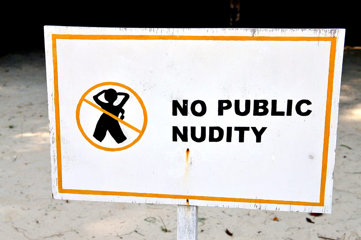 A white beach sign with a yellow border reads “NO PUBLIC NUDITY” in black capital letters. A stylized silhouette of a nude figure with hands behind the head is shown inside a circle with a diagonal slash, indicating prohibition. The background is sandy ground with some shadow and foliage.