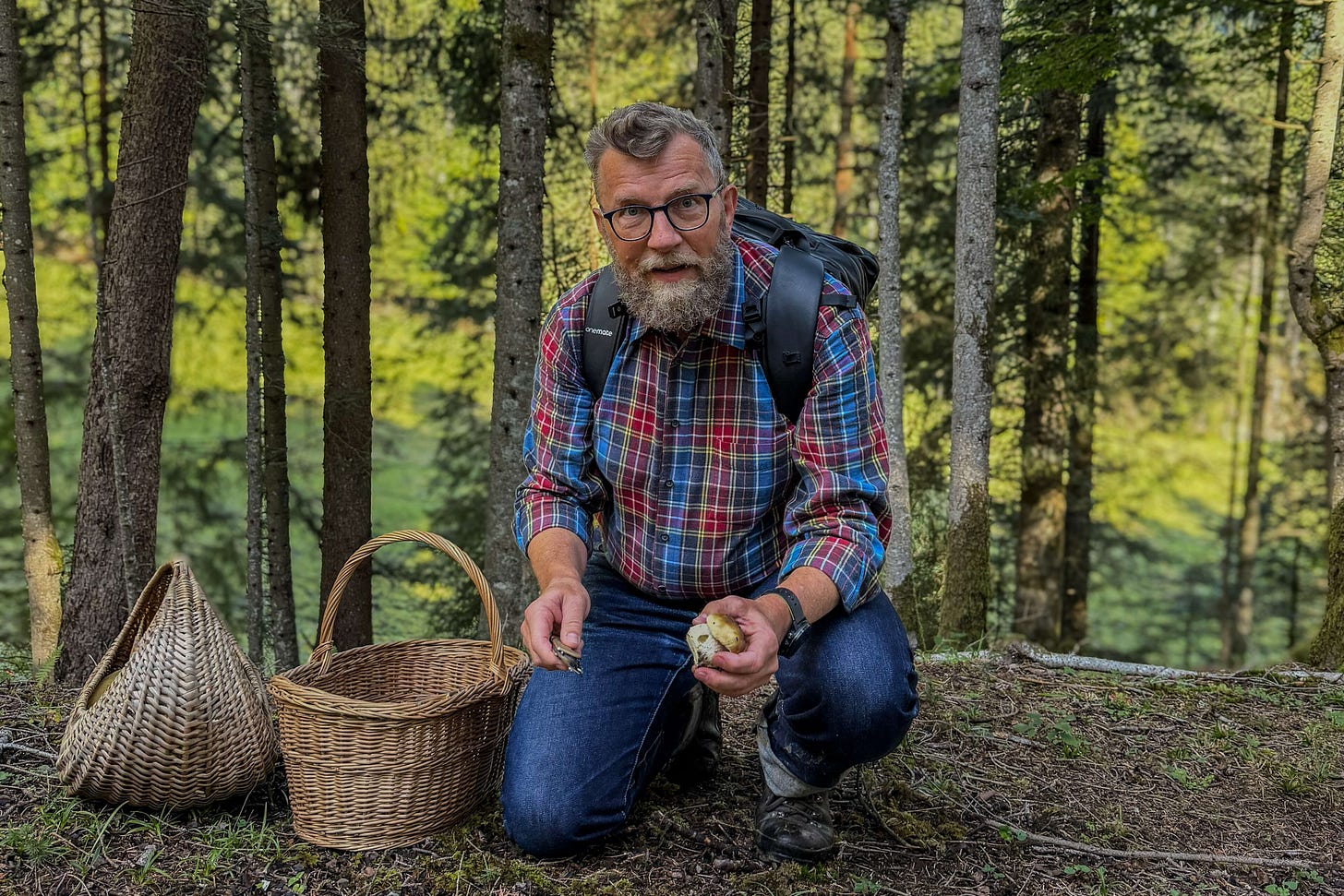 Der Agrarjournalist Jürg Vollmer – ein bärtiger Mann im karierten Hemd, Jeans und Wanderschuhen – kniet im Wald neben einem Pilzkorb. Er trägt einen Rucksack und hält einen frisch gefundenen kleinen Steinpilz in der Hand, den er direkt an der Fundstelle mit einem Messer putzt. Im Hintergrund sind Bäume und grünes Waldlicht zu sehen. Der Agrarjournalist Jürg Vollmer – ein bärtiger Mann im karierten Hemd, Jeans und Wanderschuhen – kniet im Wald neben einem Pilzkorb. Er trägt einen Rucksack und hält einen frisch gefundenen kleinen Steinpilz in der Hand, den er direkt an der Fundstelle mit einem Messer putzt. Im Hintergrund sind Bäume und grünes Waldlicht zu sehen.