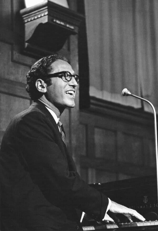 A smiling man with dark hair and glasses plays the piano while singing into a microphone.