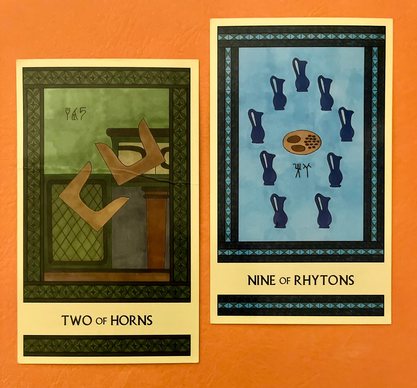 Two Minoan Tarot cards side by side on an orange surface. The Two of Horns is painted in shades of deep green and brown. It shows two pairs of tan sacred horns in midair as they tumble off a shrine. The Nine of Rhytons is painted in shades of blue. It shows nine royal blue pitchers arranged in a circle around a platter of bread and olives. 
