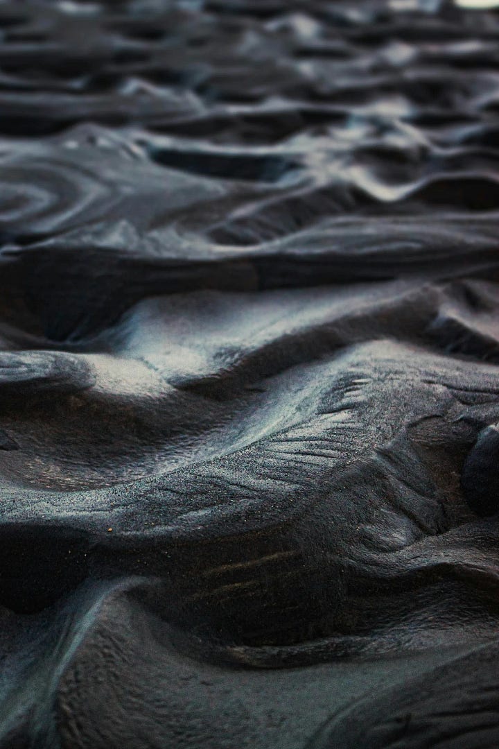 First Image: Watercolor in shades of blue, gray and black. A huge draconic silhouette approaches a pirate vessel on the sea with a curious demeanor.  Second Image: an expanse of black sands on a volcanic shore. Golden flecks are visible amidst the darkness.