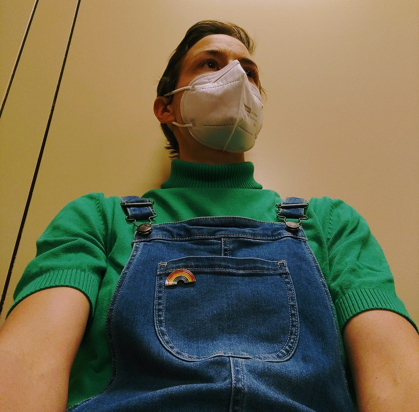A white woman with short brown hair is wearing a respiratory mask, jeans dungarees, and a mint-green shirt. She is looking to the right and is sitting in front of a beige background.