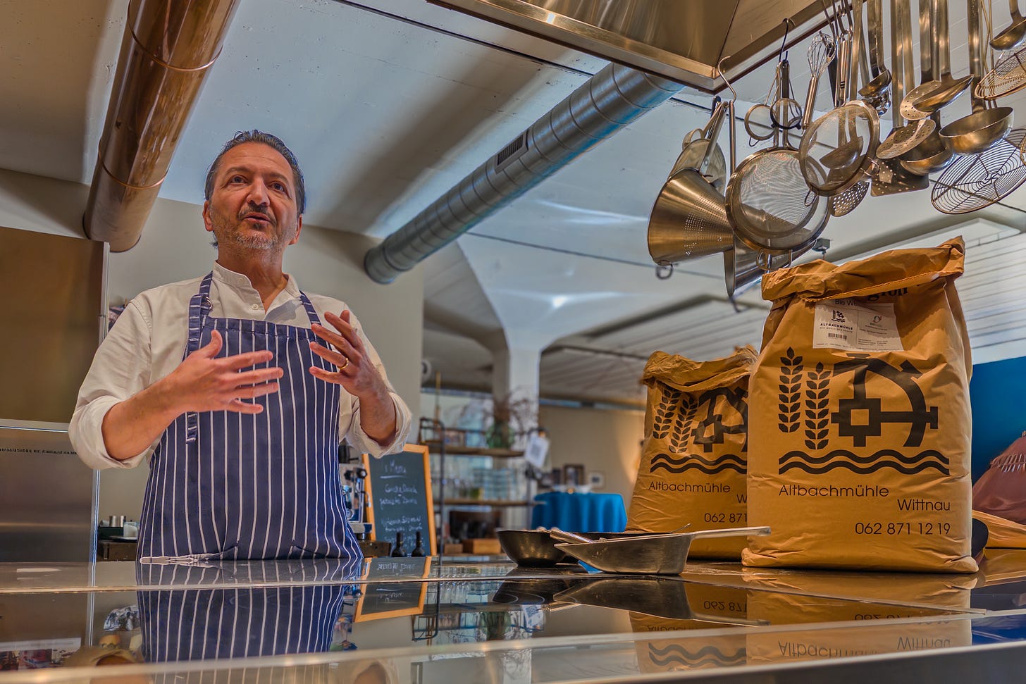 Der Schweizer Food-Journalist und Kochbuch-Autor Claudio Del Principe steht in einer Backstube in gestreifter Schürze vor zwei grossen Mehlsäcken der Altbachmühle und erklärt mit einer Handbewegung etwas. Über ihm hängen Siebe, Schöpflöffel und weiteres Küchenwerkzeug. Der Schweizer Food-Journalist und Kochbuch-Autor Claudio Del Principe steht in einer Backstube in gestreifter Schürze vor zwei grossen Mehlsäcken der Altbachmühle und erklärt mit einer Handbewegung etwas. Über ihm hängen Siebe, Schöpflöffel und weiteres Küchenwerkzeug.