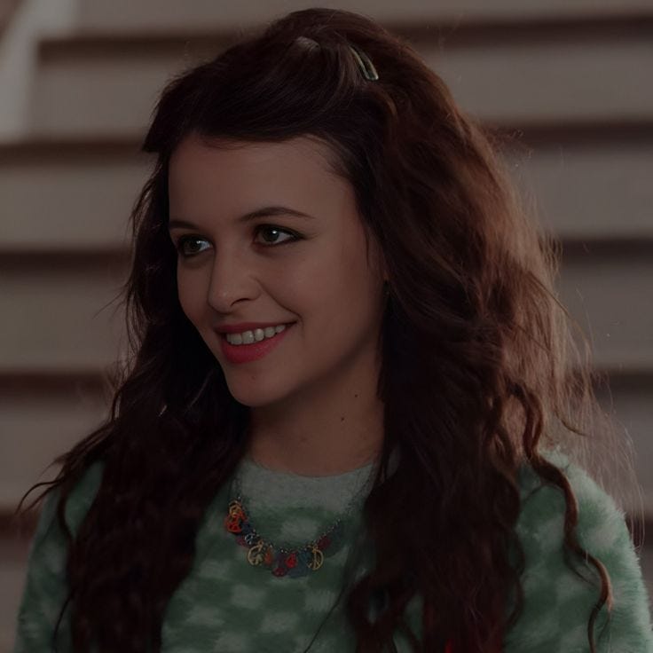 This may contain: a woman with long brown hair wearing a green sweater and smiling at the camera while standing in front of stairs