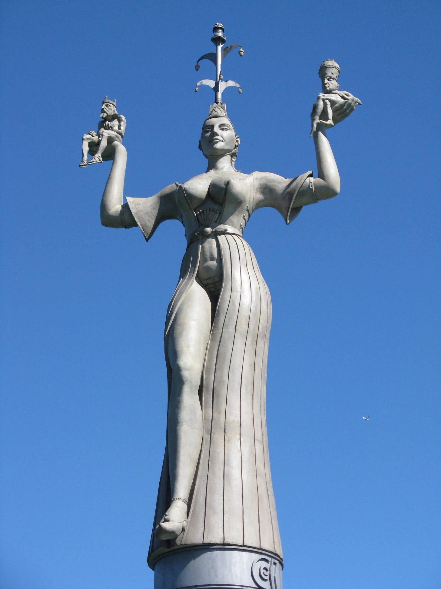Against blue sky, concrete statue of woman in close-fitting robe partially open at chest and legs stands holding two much smaller seated men in her upheld palms