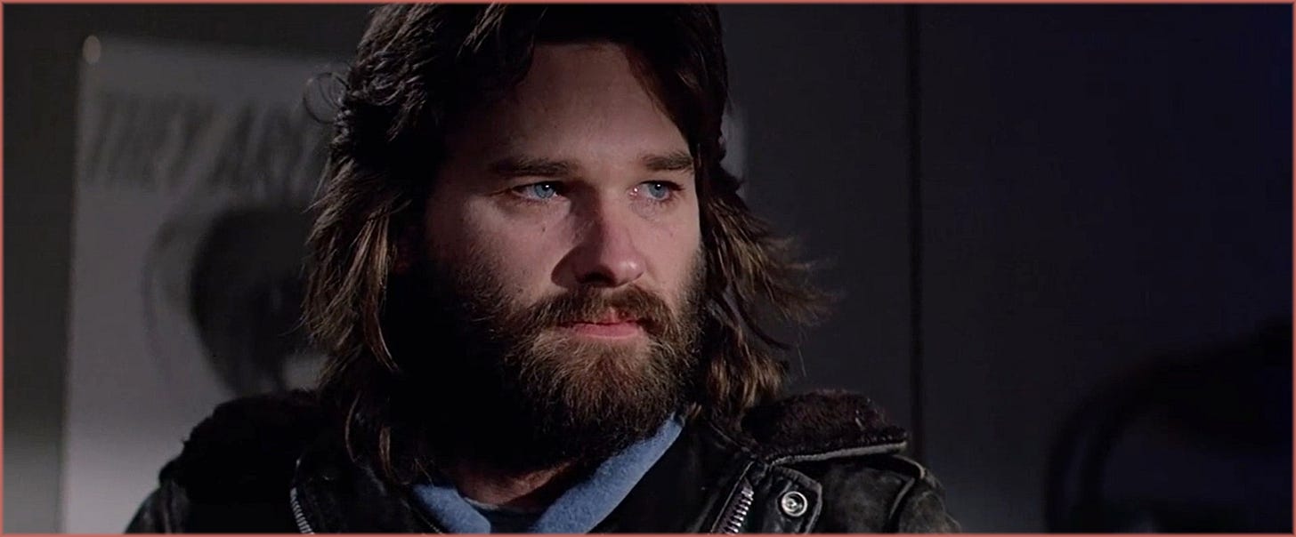 Close-up from The Thing (1982): MacReady (Kurt Russell) during the blood test sequence, looking off-camera with calm, calculating focus. Long hair and beard grown over nearly a year of pre-production.