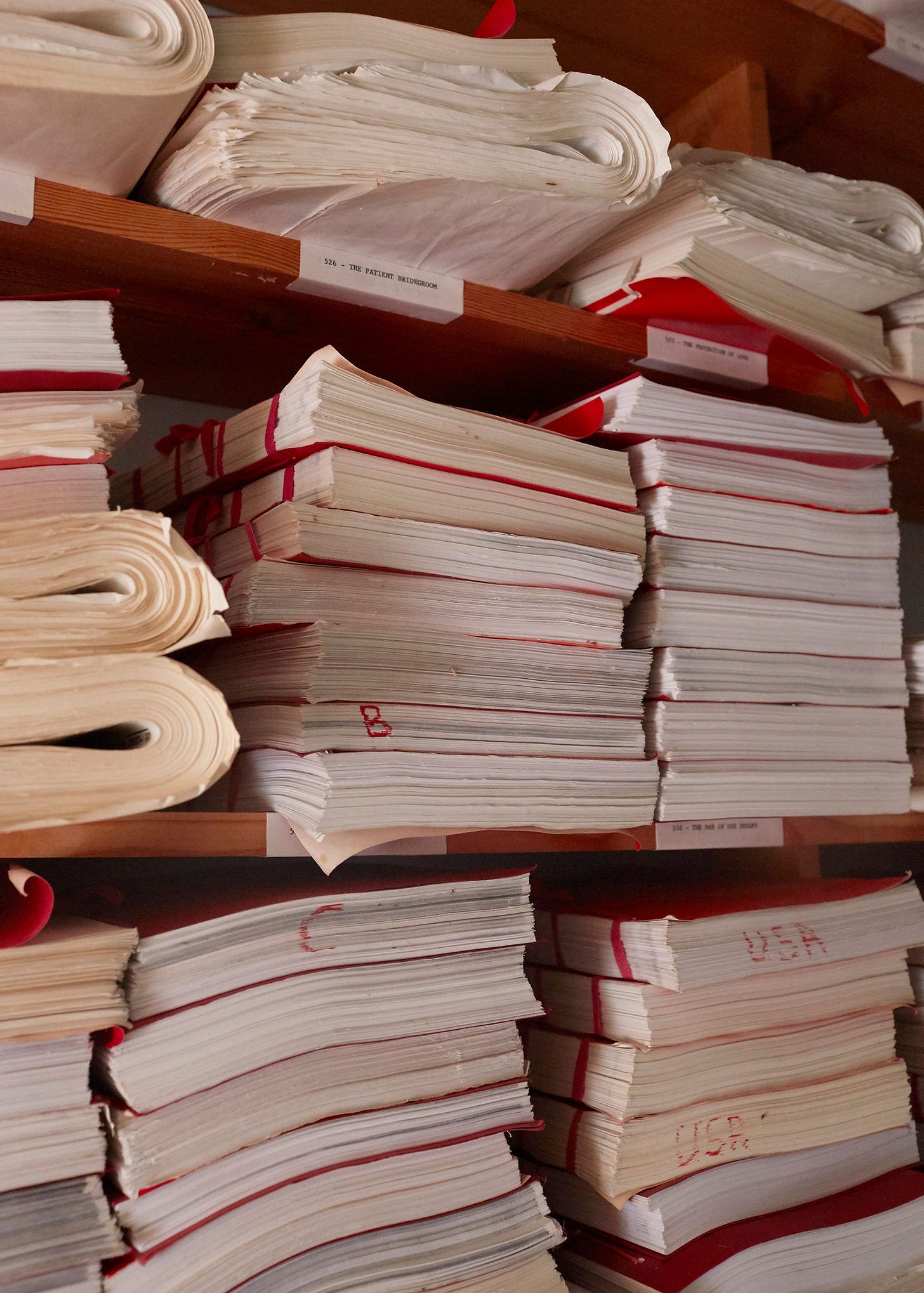 Stacks of files on wooden shelves with red labels and visible text like "USA." Papers are worn and slightly curled. Archive setting. Barbara Cartland Claudia Legge