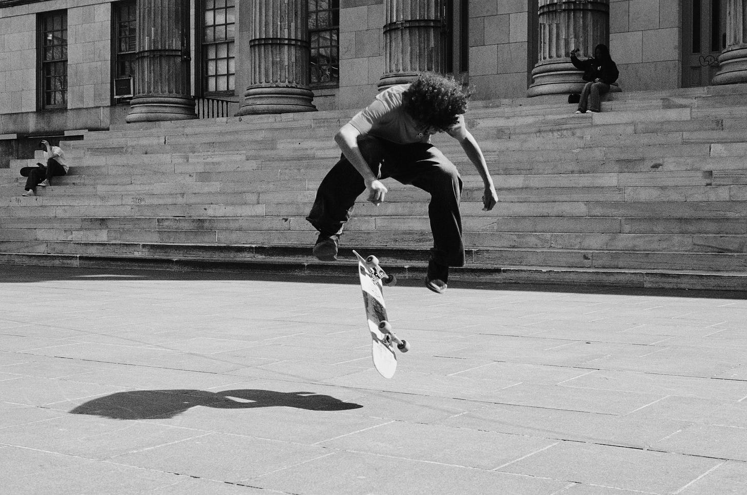 A teenage boy skateboarding is suspended in the air while doing a trick in a public plaza. A girl sits behind him on the steps taking a selfie on her phone.