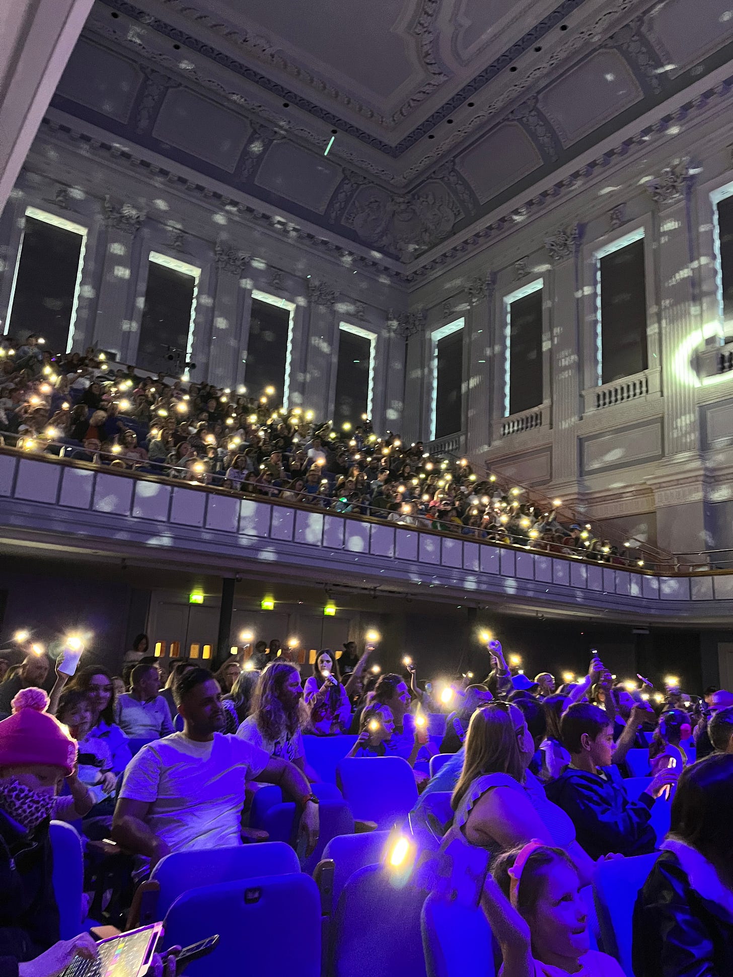 A photo of a packed concert hall at a rock gig. The audience are holding the torches on their phones to create a stunning effect of a sea of lights in a darkened room.