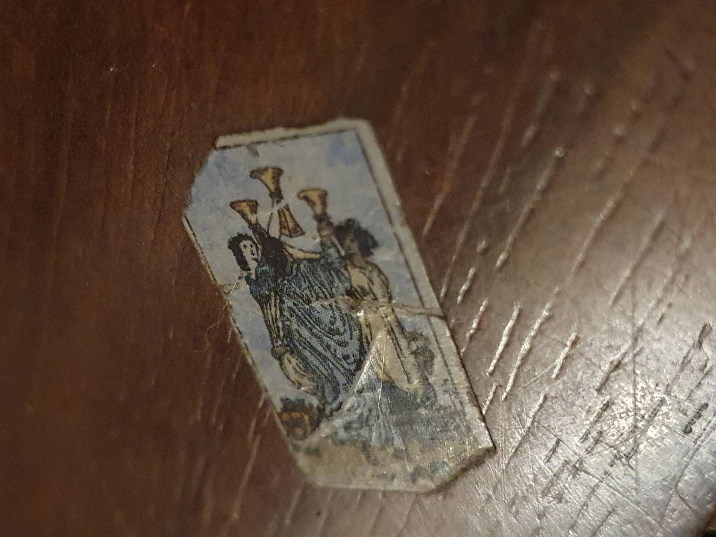 A tiny, torn and weathered fragment of a traditional Three of Cups tarot card, showing three figures raising golden cups, resting on a scratched wooden surface. A tiny, torn and weathered fragment of a traditional Three of Cups tarot card, showing three figures raising golden cups, resting on a scratched wooden surface.