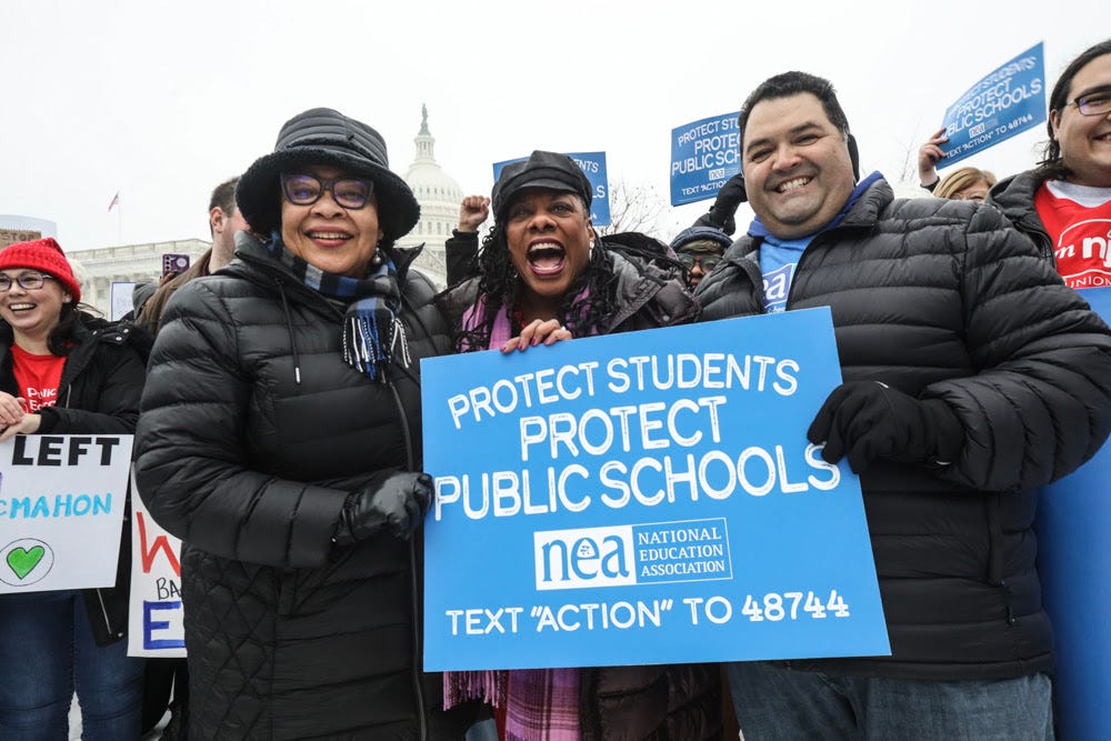 Princess Moss, Becky Pringle, and Noel Candelaria stand outside in winter clothing, along with a diverse group of NEA members, holding signs that say, ''protect students protect public schools.'' Princess Moss, Becky Pringle, and Noel Candelaria stand outside in winter clothing, along with a diverse group of NEA members, holding signs that say, ''protect students protect public schools.''