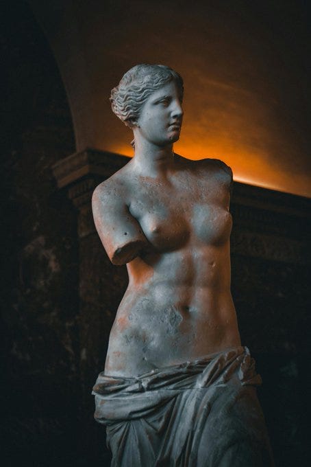 Marble statue of Venus de Milo standing with missing arms, exposed breasts and defined abdominal muscles visible on the upper body, draped fabric covering the lower body and legs, set in a dimly lit arched museum interior with stone architecture in the background. Marble statue of Venus de Milo standing with missing arms, exposed breasts and defined abdominal muscles visible on the upper body, draped fabric covering the lower body and legs, set in a dimly lit arched museum interior with stone architecture in the background.