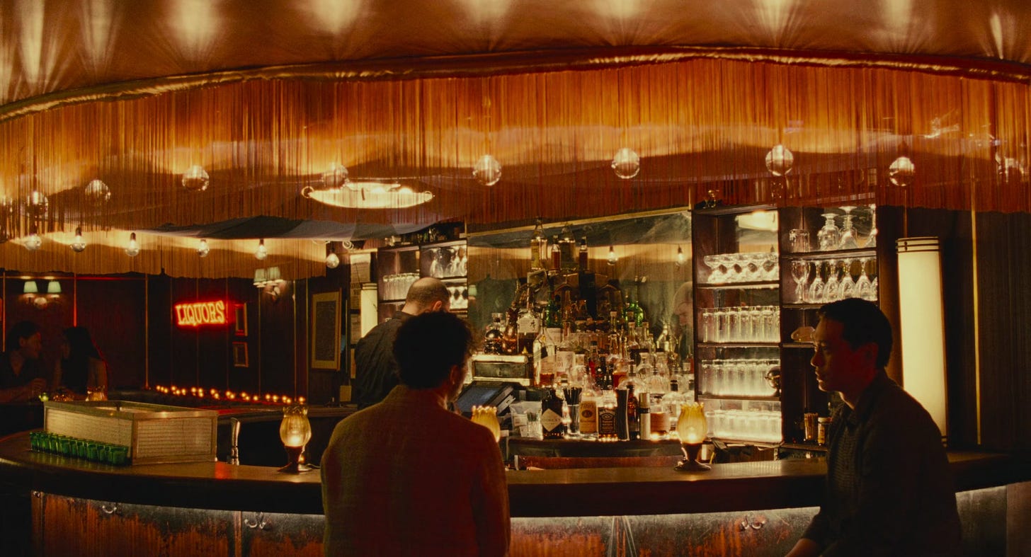 Vista do bar onde Nora, Hae Sung e Arthur se encontram. Os dois personagens são vistos sentados juntos no balcão iluminado por luzes amareladas, criando uma atmosfera íntima e nostálgica.