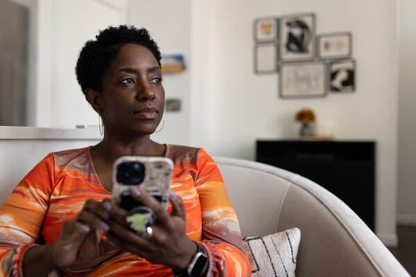 Tia-Maria Smith sits in a chair against pillows wearing an orange top and holding a phone.