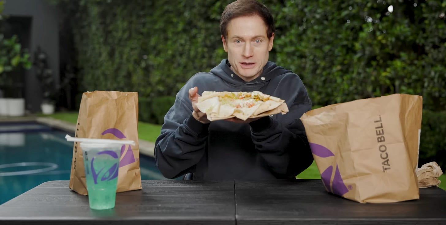 Bryan holding up an open Crunchwrap; he's at a table surrounded by Taco Bell bags and a Baja Blast; his backyard is in the background Bryan holding up an open Crunchwrap; he's at a table surrounded by Taco Bell bags and a Baja Blast; his backyard is in the background