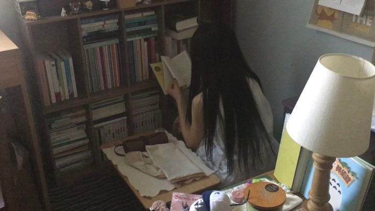 This may contain: a woman with long hair sitting in front of a book shelf next to a lamp