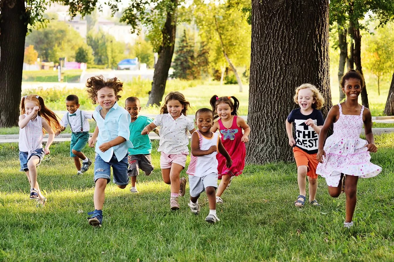 Group of smiling young children of diverse backgrounds running together through a sunlit grassy park with trees. Group of smiling young children of diverse backgrounds running together through a sunlit grassy park with trees.