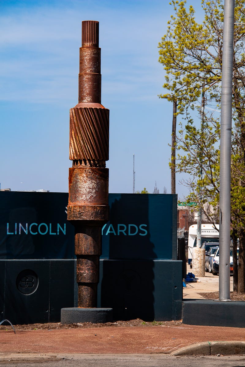Tall rusted industrial gear-like sculpture standing in front of the Lincoln Yards signage with a city street in the background.