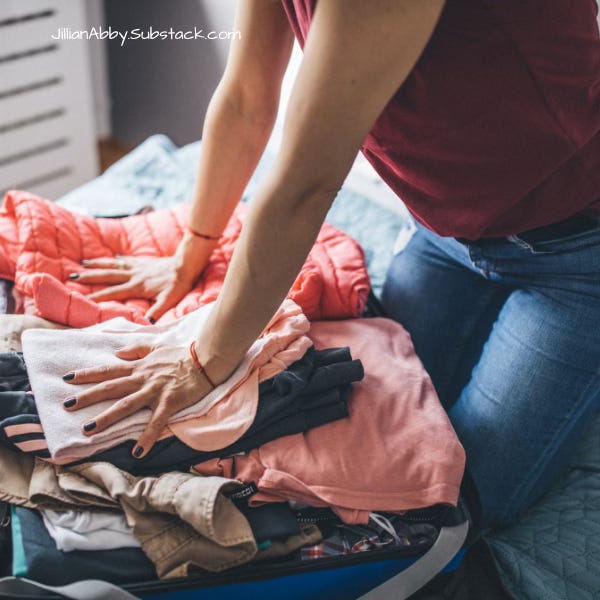 A person in a maroon sleeveless top and jeans is packing a suitcase, pressing down on a pile of folded clothes including a pink jacket and various shirts to make them fit. The photo is taken indoors, and the website "JillianAbby.Substack.com" is written in the upper left corner.