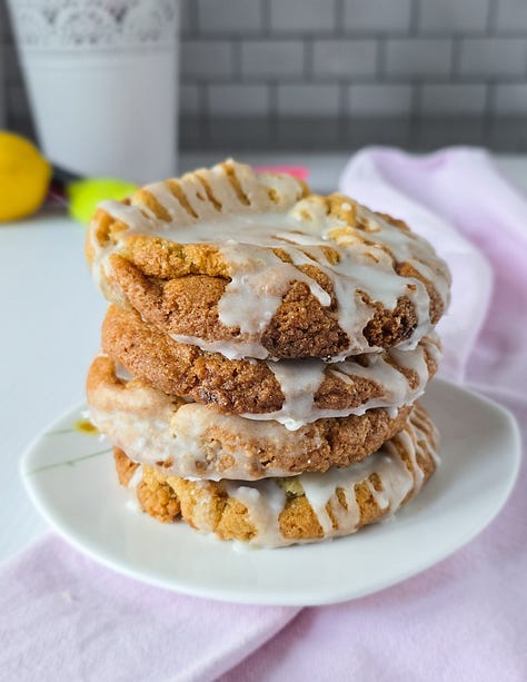 a pink baking tray with almond macaroons and glace cherries on it. A cooling rack with apple and blackberry muffins on it with apples at the top right hand of the shot. A pink cake plate with a 4 layer spiced plum upside down cake on it.