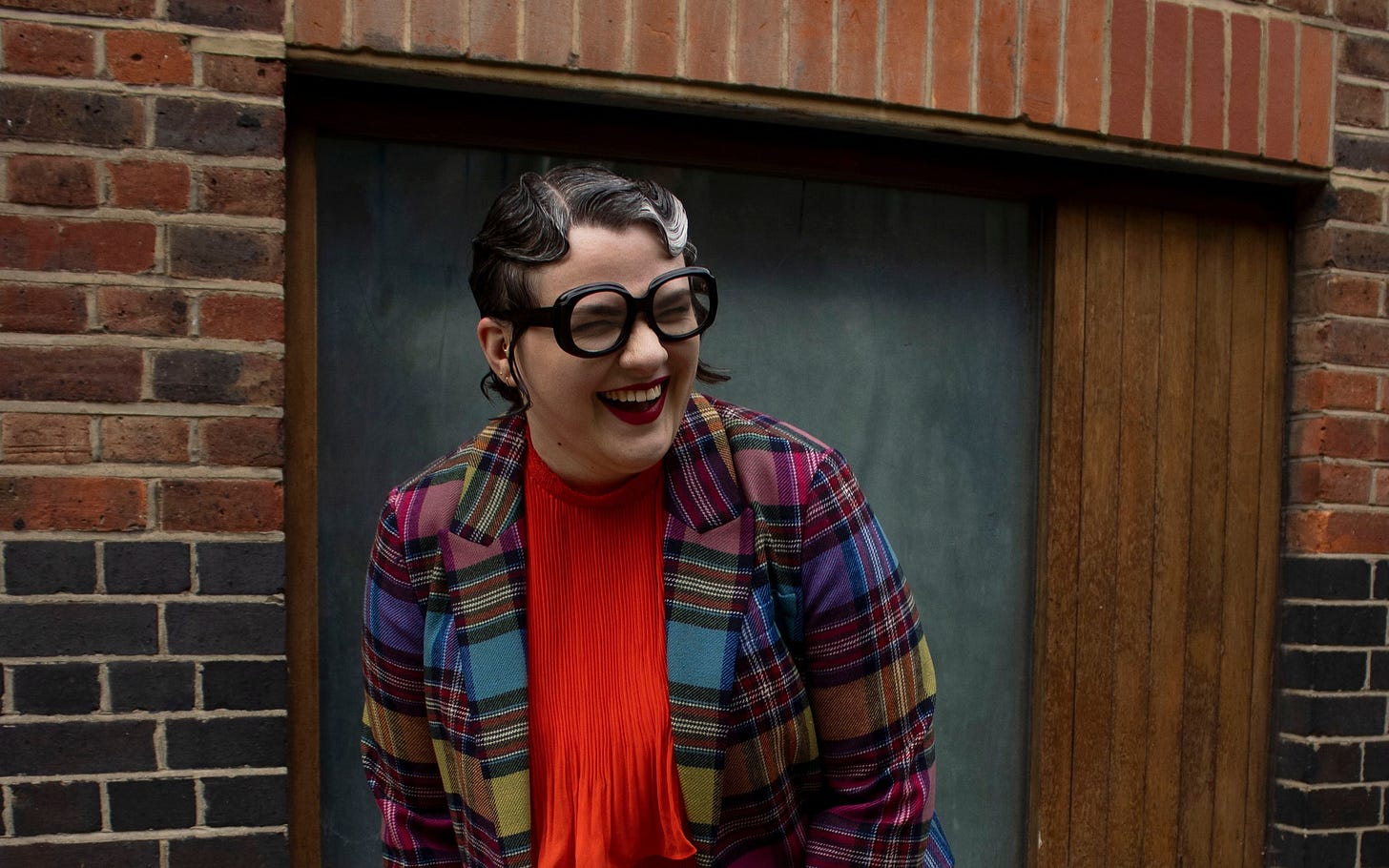 Jennifer Heinen laughing, wearing red lipstick, round black glasses, and a multicoloured tartan blazer over an orange top.