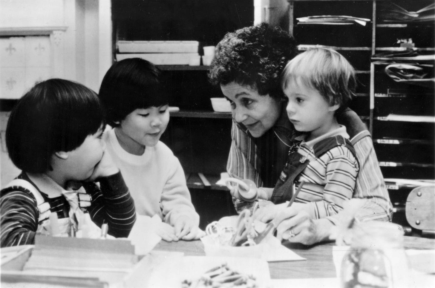 Black and white classroom photograph showing Vivian Gussin Paley, with curly dark hair, leaning in to talk with three young children at a table. The children, who appear to be of different ethnicities, are focused on toys or materials in front of them. Shelves with books and supplies are visible in the background.