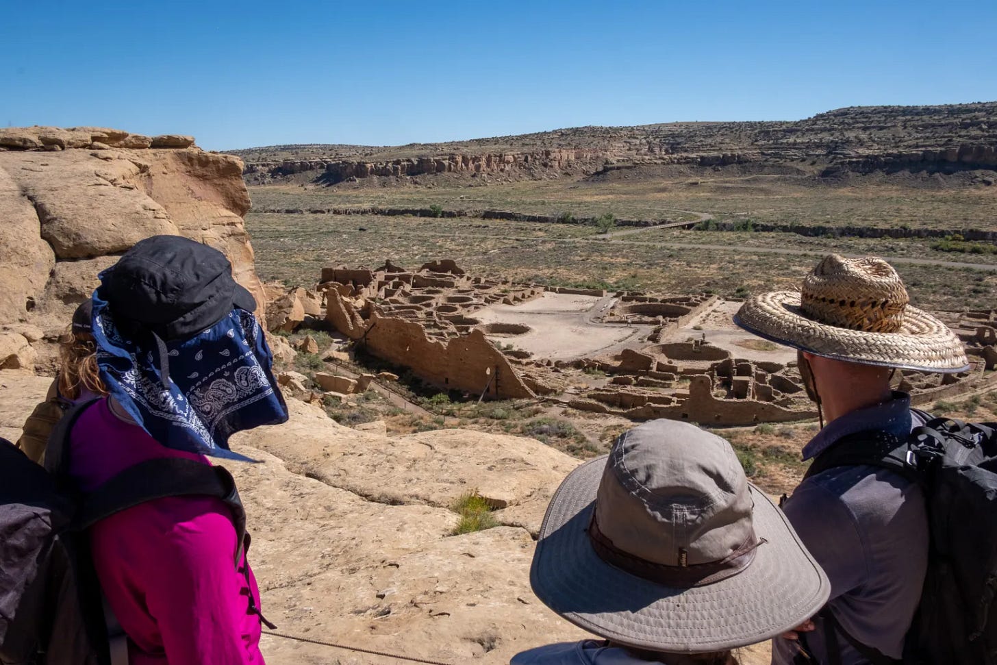 Three people wearing hats and sun protection peer across a rock formation toward an ancient structure in the valley below. Three people wearing hats and sun protection peer across a rock formation toward an ancient structure in the valley below.