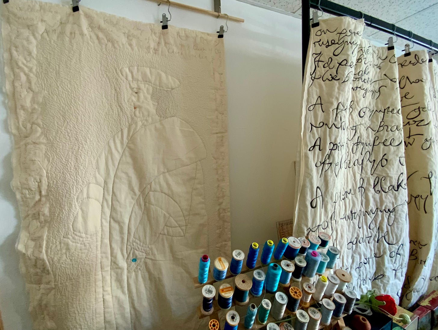 A large stitched portrait of Thomas Cromwell hangs on a wall behind a rack of colourful thread. A large quilted piece showing sixteenth century handwriting is next to Cromwell. Some desk space is showing beneath that.