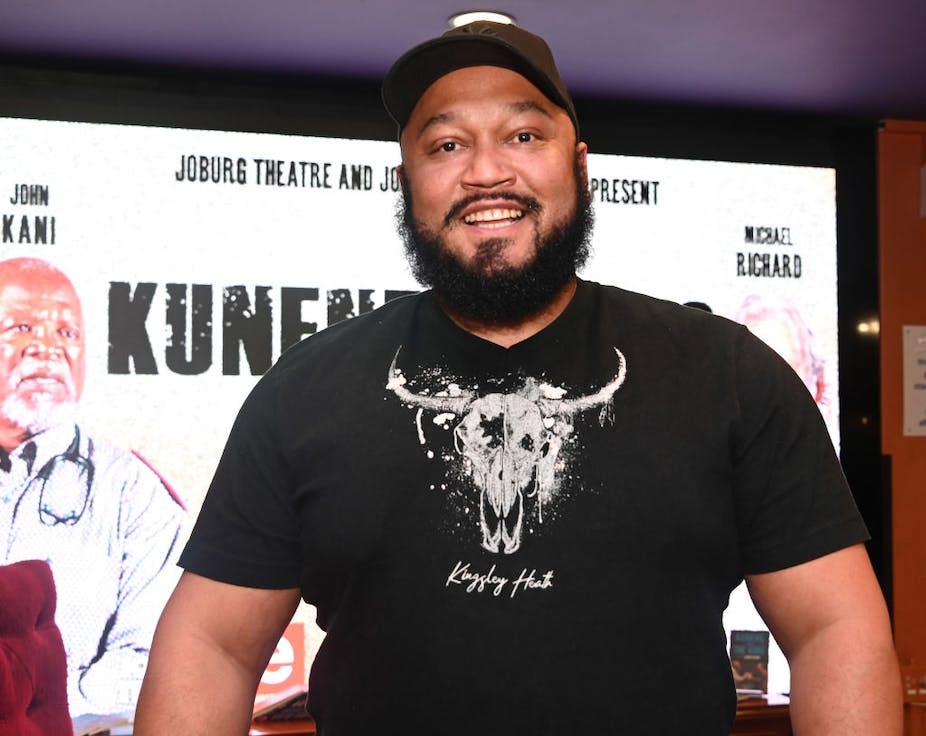 A man with a beard in a cap and T-shirt smiles as he looks at the camera, a theatre banner behind him.
