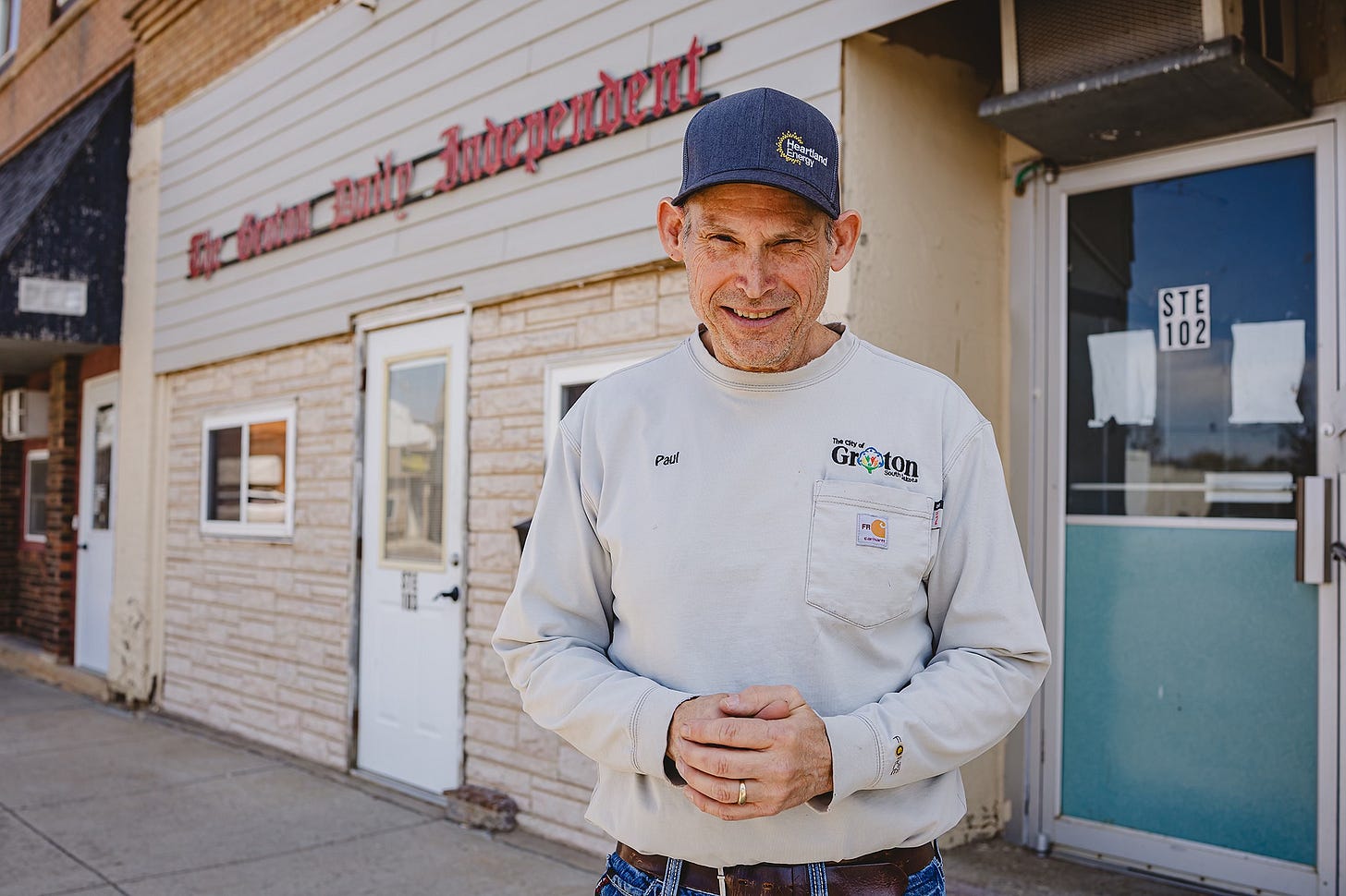 Paul Kosel, known as "Paper Paul," stands smiling in front of the Groton Daily Independent office on Main Street. He wears a grey long-sleeve "City of Groton" work shirt and a "Heartland Energy" baseball cap, visually representing his dual roles as a local news publisher and a municipal employee in rural South Dakota. Paul Kosel, known as "Paper Paul," stands smiling in front of the Groton Daily Independent office on Main Street. He wears a grey long-sleeve "City of Groton" work shirt and a "Heartland Energy" baseball cap, visually representing his dual roles as a local news publisher and a municipal employee in rural South Dakota.