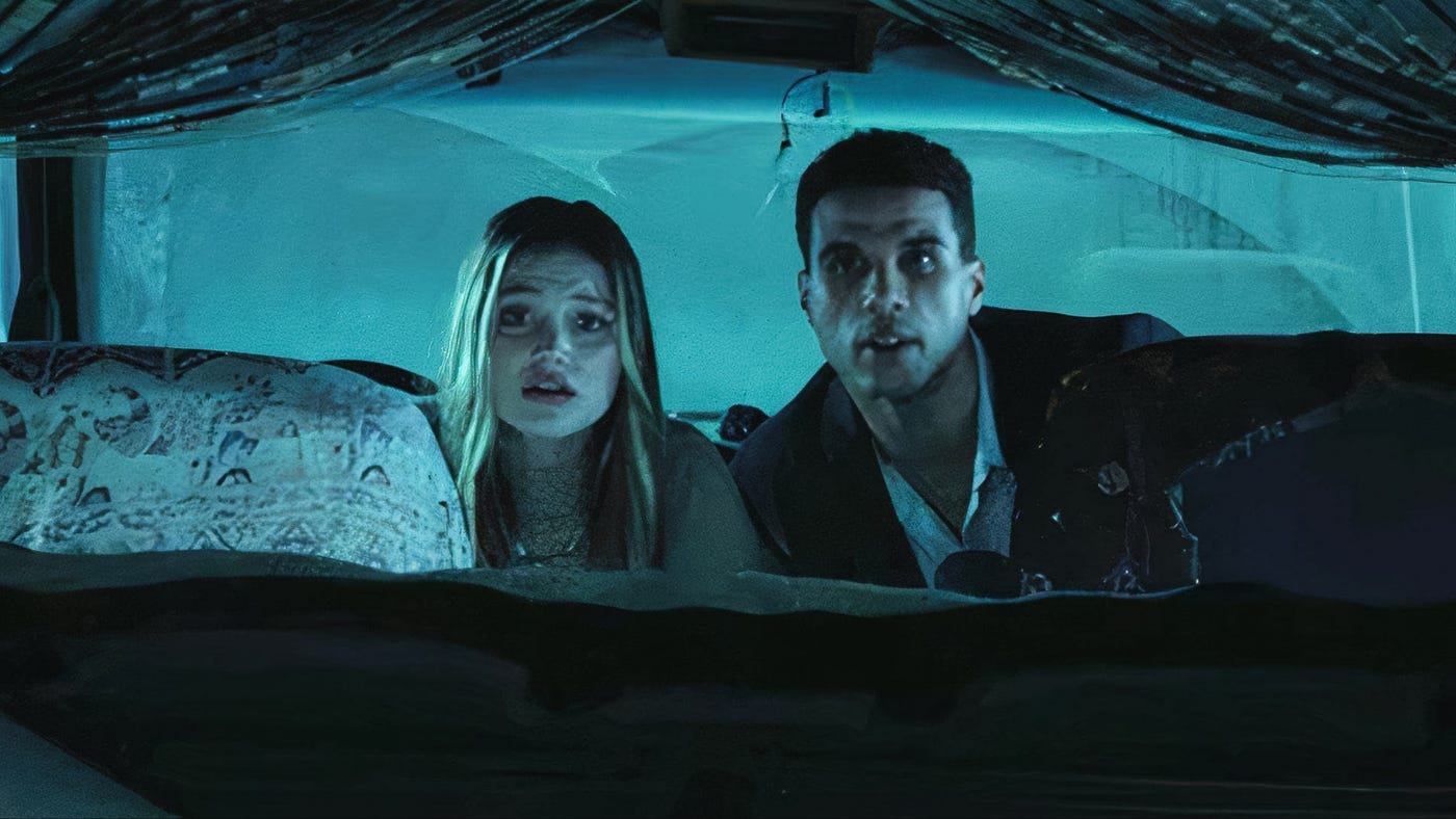 A man and woman sit frozen in fear inside a dimly lit van with a shattered windshield and teal lighting.