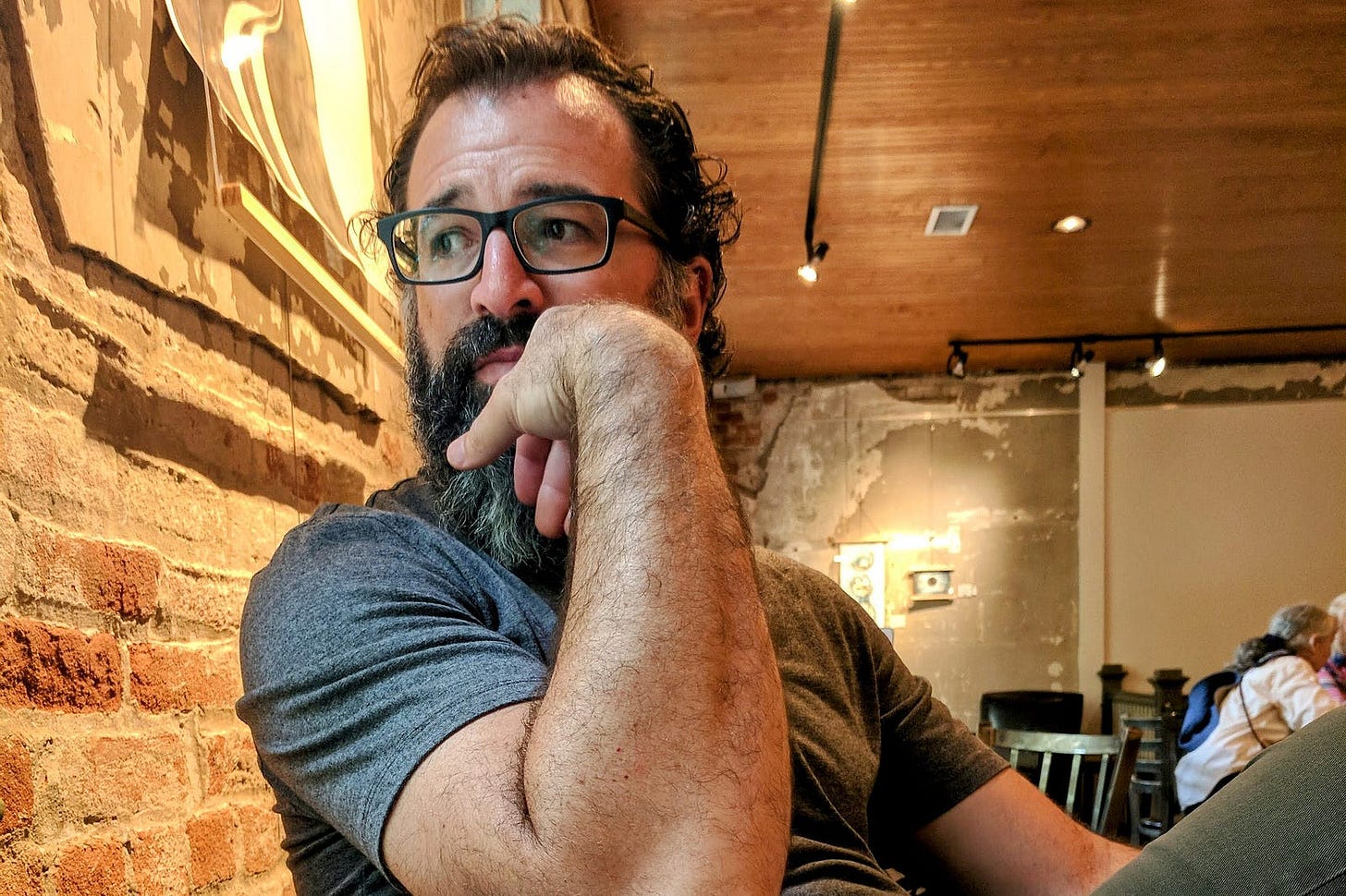 The author with dark curly hair, glasses, and a salt-and-pepper beard sits indoors against an exposed brick wall. He is wearing a grey t-shirt and resting his chin thoughtfully on his hand, looking off to the side. The warm-toned room has a wooden ceiling and worn plaster walls, with other people seated in the background. The author with dark curly hair, glasses, and a salt-and-pepper beard sits indoors against an exposed brick wall. He is wearing a grey t-shirt and resting his chin thoughtfully on his hand, looking off to the side. The warm-toned room has a wooden ceiling and worn plaster walls, with other people seated in the background.