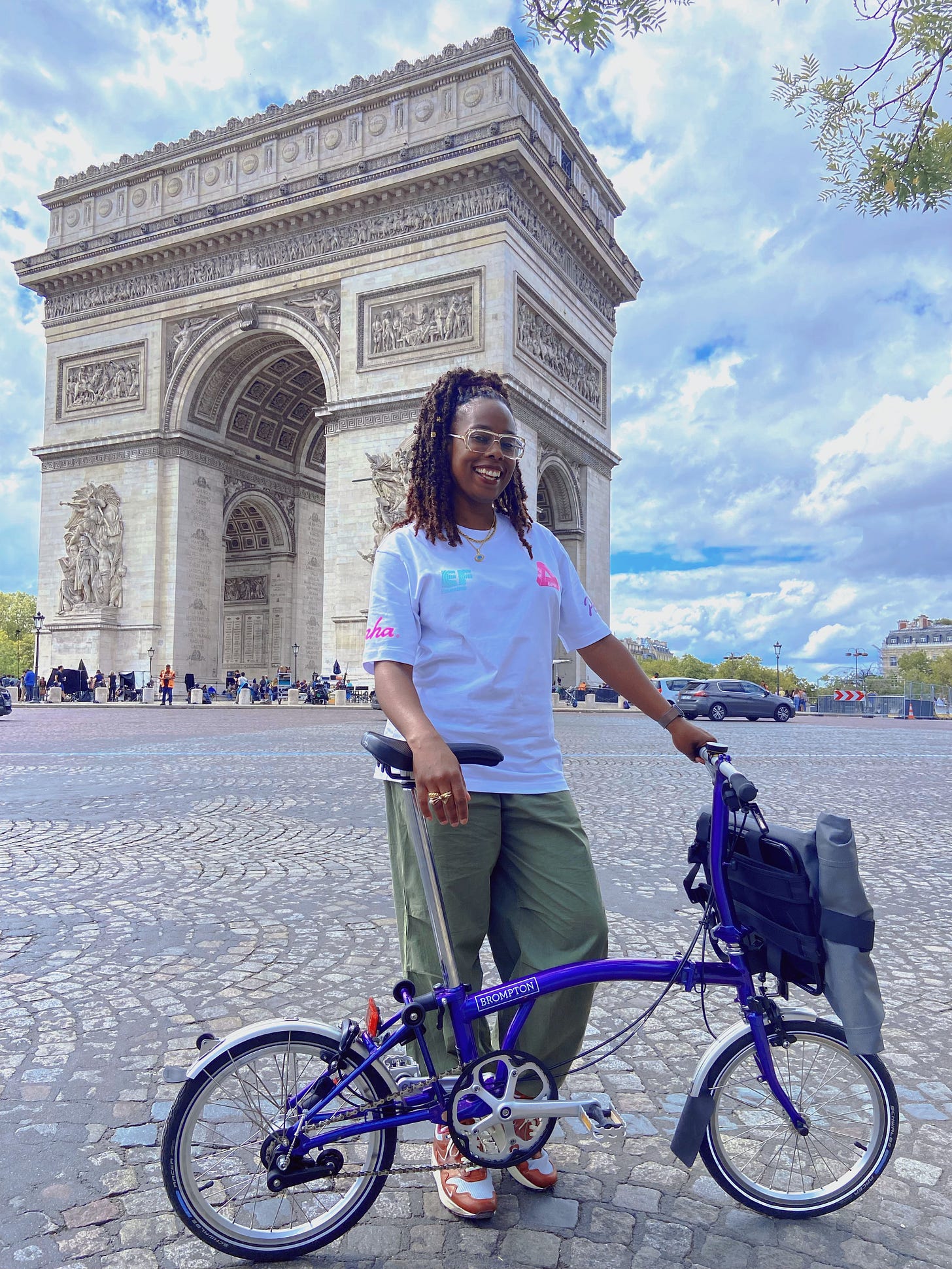 Jools is standing in front of the arc de triomphe in Paris, holding her purple brompton bicycle. She is wearing a white t-shirt and green trousers and smiling. Jools is standing in front of the arc de triomphe in Paris, holding her purple brompton bicycle. She is wearing a white t-shirt and green trousers and smiling.