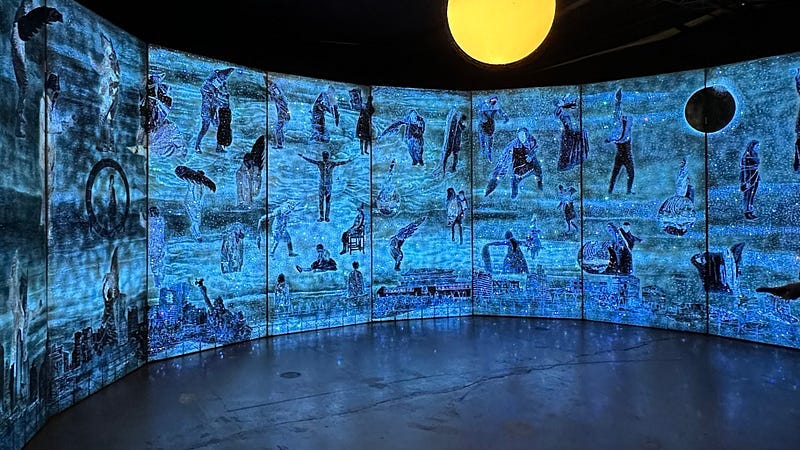 A series of tall, canvased panels form a panoramic arc. They are illuminated with a marine blue light. Comprised of individual figures and monuments, each panel displays a different depiction of destruction or decay. One figure wears a gas mask; the Statue of Liberty appears to have crumbled, with only its upper half visible and careening to the side.