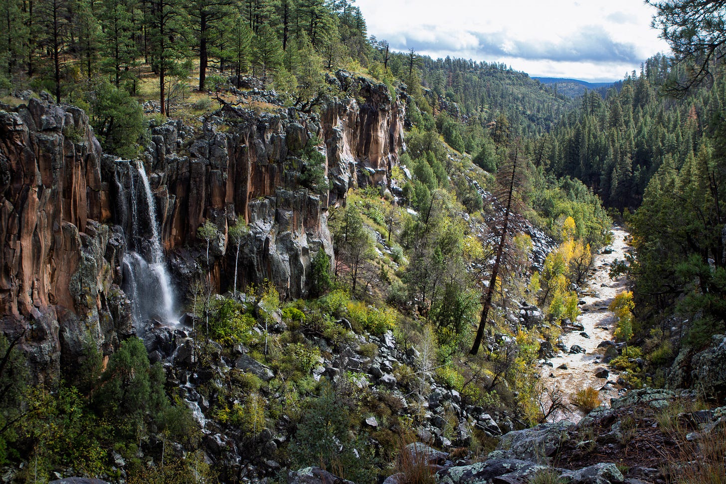 dramatic waterfall down an orange/brown cliff to a dark green pine filled valley with a stream running through it. dramatic waterfall down an orange/brown cliff to a dark green pine filled valley with a stream running through it.