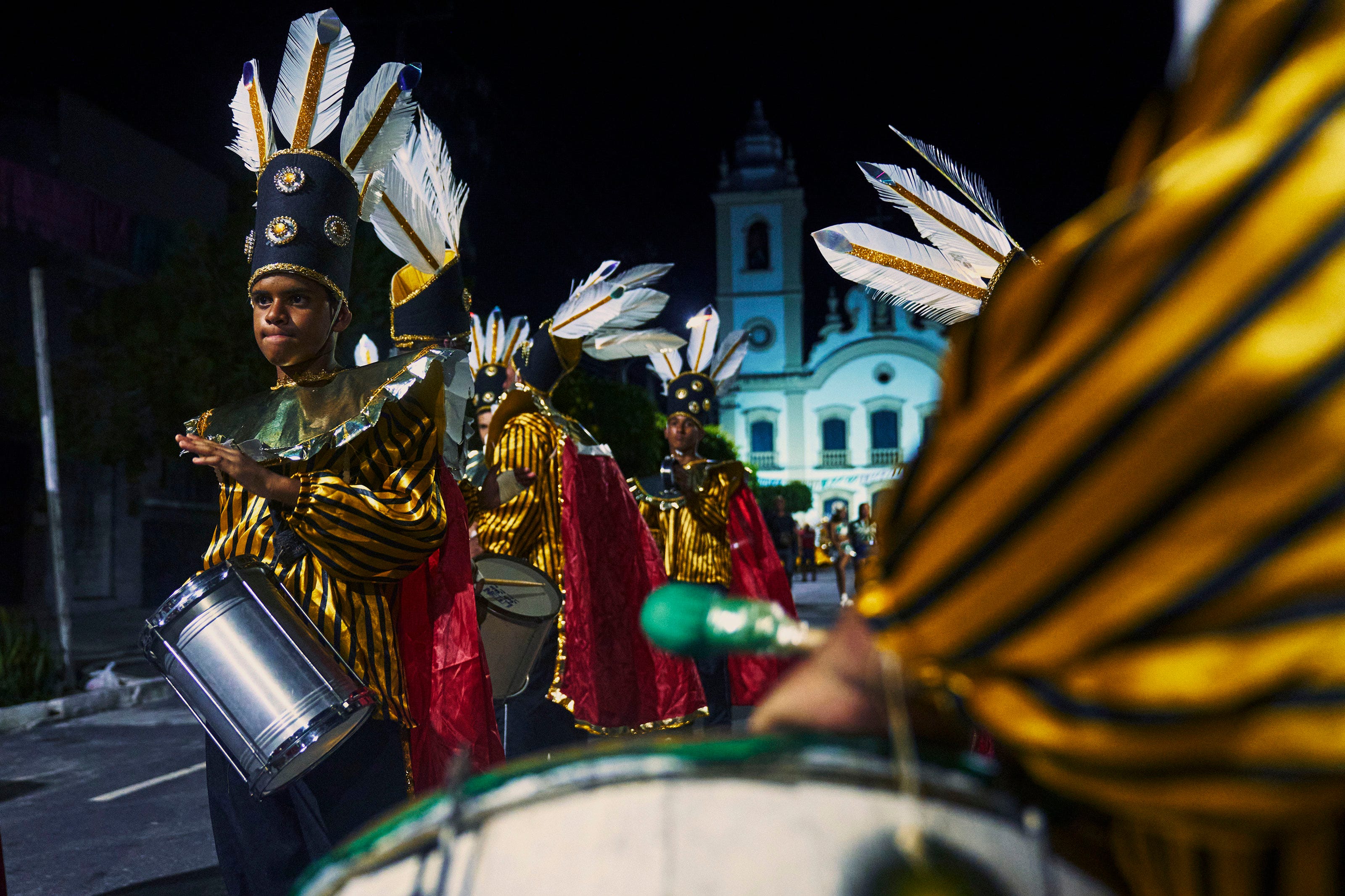 People dressed in vibrant striped costumes with feathered headdresses play drums during a lively nighttime street parade, with a church visible in the background.