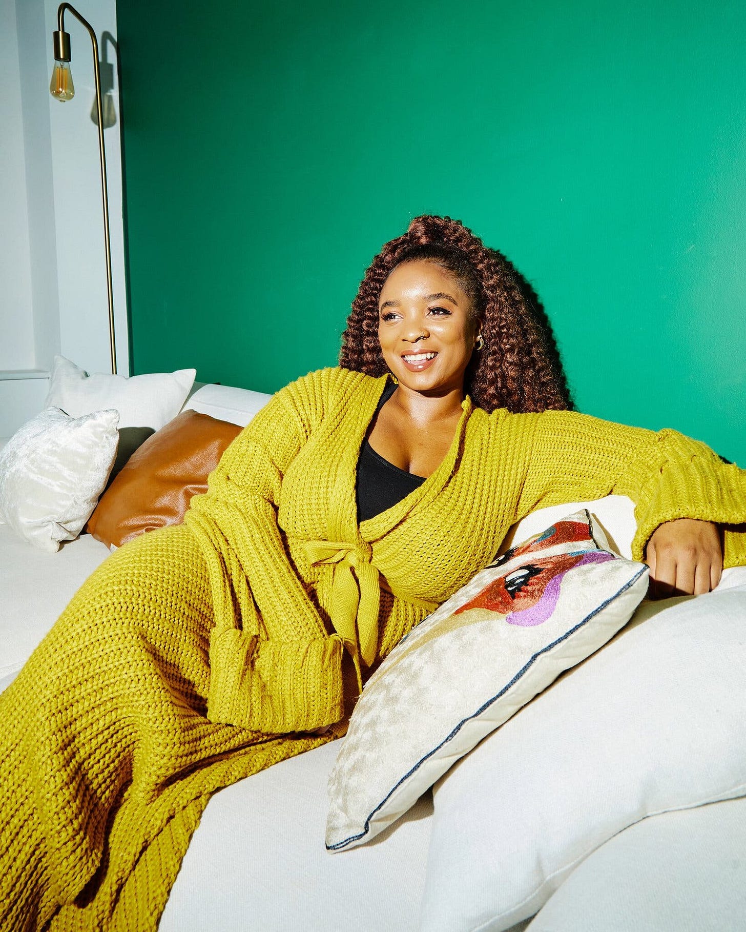 Tiffany Chanel Knighten sitting on a white couch in front of a green wall. She is dressed in a knit yellow sweater dress. 