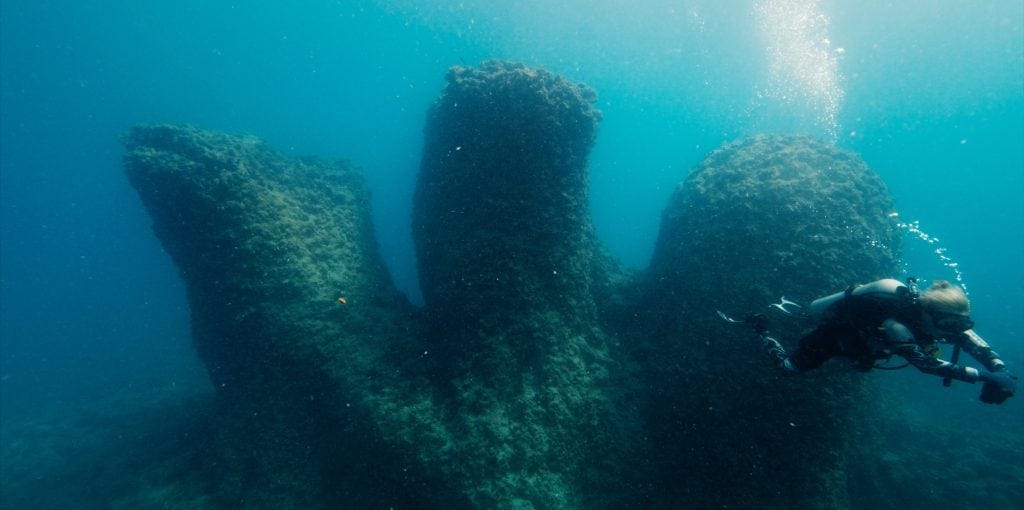 Scuba diver exploring underwater ruins resembling giant stone columns from ancient Alexandria, linked to Cleopatra’s sunken palace in the Mediterranean Sea.
