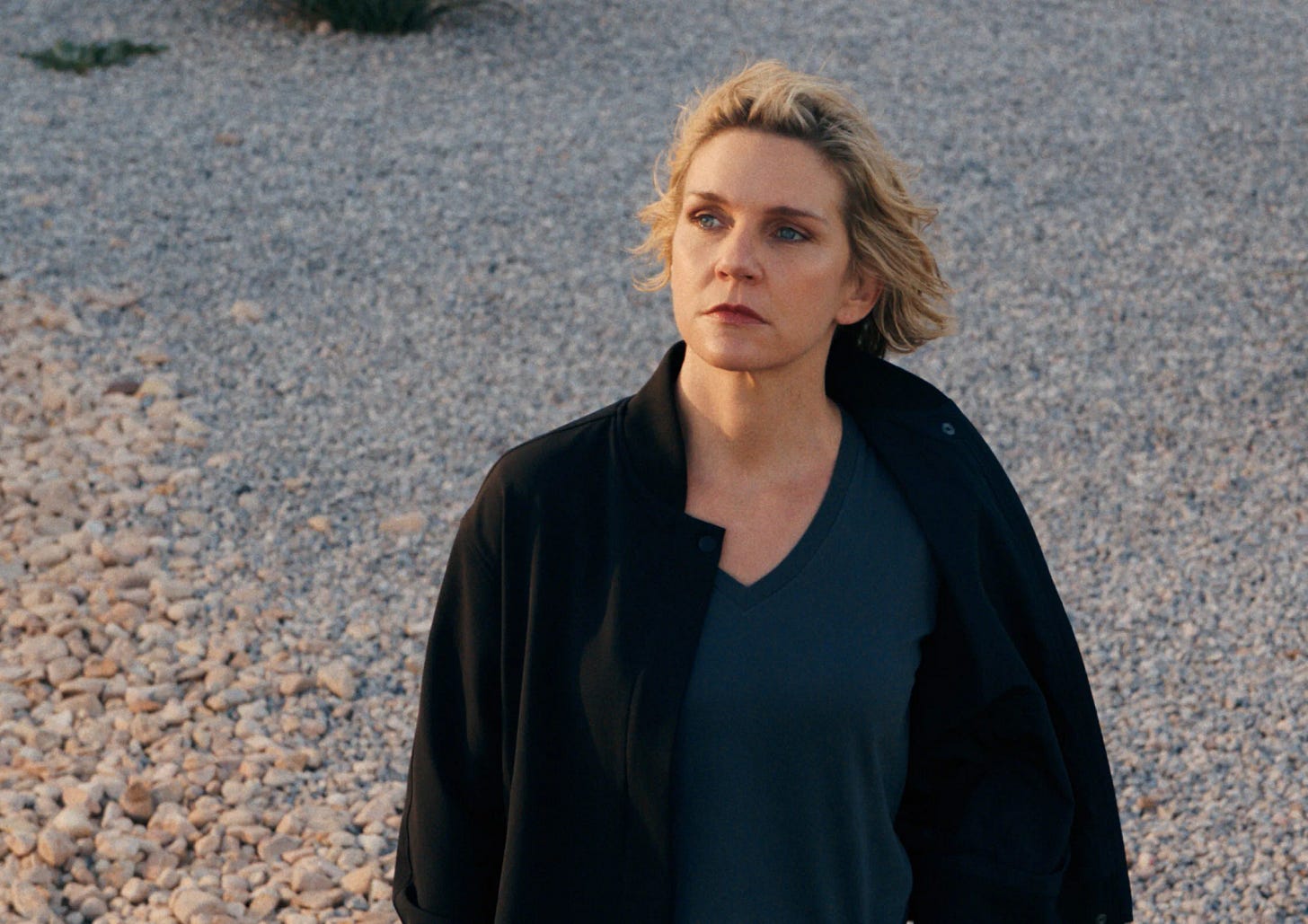 Carol, blond white woman, stands on rocks looking at sky; she is wearing a black coat
