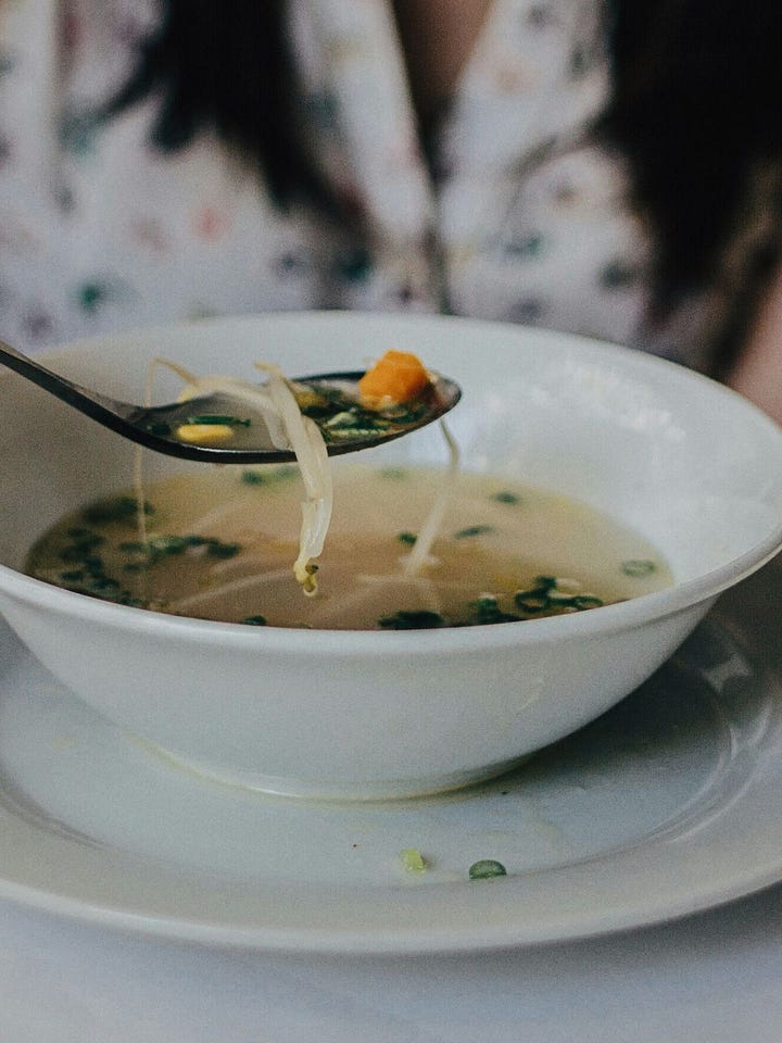 Soft domestic warmth during Imbolc: a wood-burning stove with stacked logs beside it, and a simple bowl of clear nourishing soup held mid-spoon, evoking heat, sustenance, and quiet restoration.